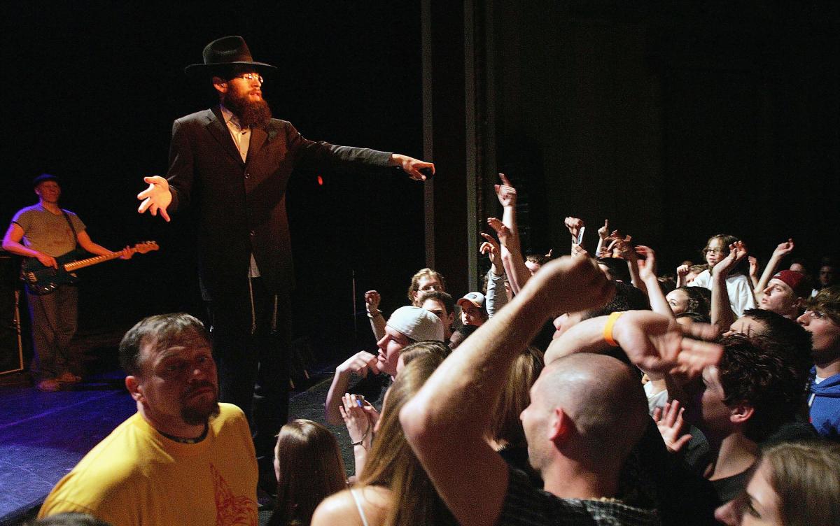 Matisyahu on stage, in a black suit and black hat, points to a crowd of concert goers while performing