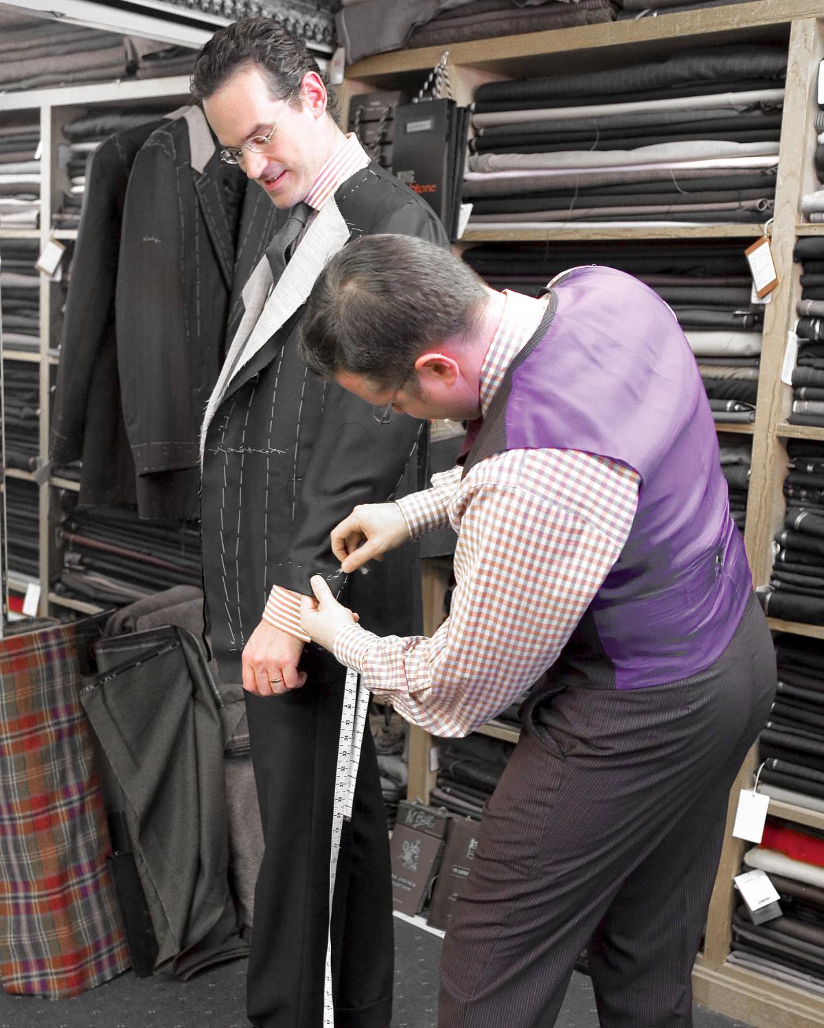 A tailor in a purple vest measures the cuff on Anton's suit jacket