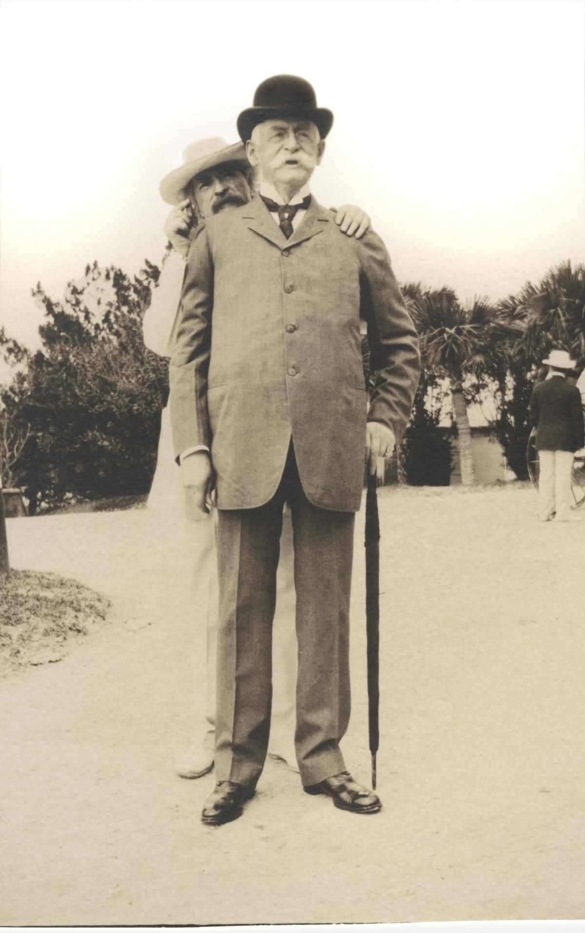 One man stands behind the other, holding onto his shoulders, both dressed in light colored suits and hats