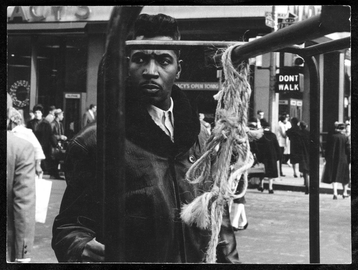 A well-dressed Black man with hair slicked back, wearing a winter leather coat and pushing a garment trolley