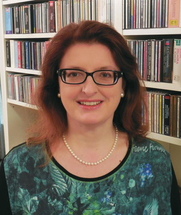 Fauser, wearing rectangular glasses and a green patterned top, standing in front of a full bookshelf