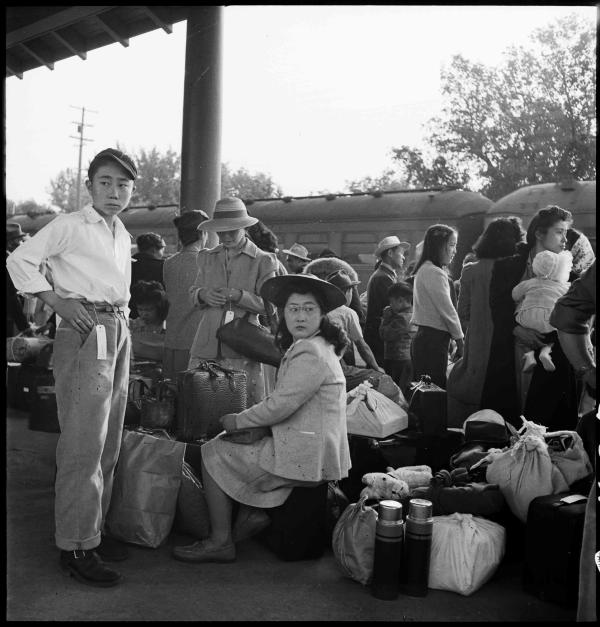 Black-and-white photo of Japanese Americans crowded at bus station with their suitcases, waiting for transport to an internment camp