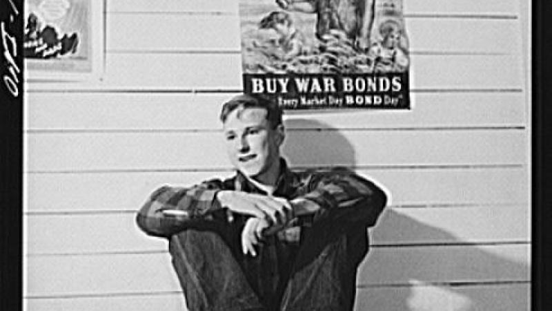 Black and white photo of young man sitting on table, war poster behind him